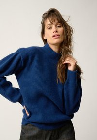 Niebieski sweter robiony na drutach z wysokim kołnierzem, z prążkowaną teksturą i lekko bufiastymi rękawami, zestawiony z ciemnymi dżinsami.