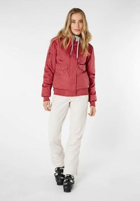 Chaqueta bomber roja con dos bolsillos frontales, forro interior gris, puños y base de canalé. Combinada con pantalones blancos y botas de esquí negras.
