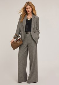 Grijze geruite dubbel-gebreide blazer gecombineerd met wijde broekspijpen, zwart topje en een bruine handtas. Bevat een geometrische ketting.