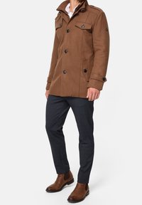 Bruine jas van wolmix met dubbel rij knopen, twee voorzakken, schouderepauletten en zwarte knopen, gecombineerd met donkere broek en bruine laarzen.