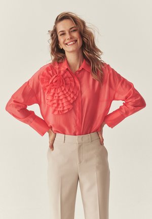 Uśmiechnięta kobieta o falowanych blond włosach, ubrana w koralową bluzkę z dużym falbanowym detalem oraz beżowe spodnie z wysokim stanem, z rękami na biodrach.