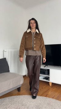 Casaco de camurça castanho com gola branca em pelo sintético, usado sobre um suéter cinzento de malha, combinado com calças largas castanhas em imitação de pele.