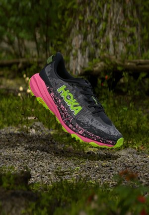 Schwarzer und grauer Hoka-Laufschuh mit neongrünem Logo und Sohle, pinker Zwischensohle, dargestellt in der Luft über einem Kiesweg in einer Waldumgebung.