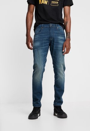 Mann trägt ausgewaschene blaue Slim-Fit-Jeans, schwarze Sneaker und ein schwarzes T-Shirt mit gelbem Text vor hellem, einfarbigem Hintergrund.