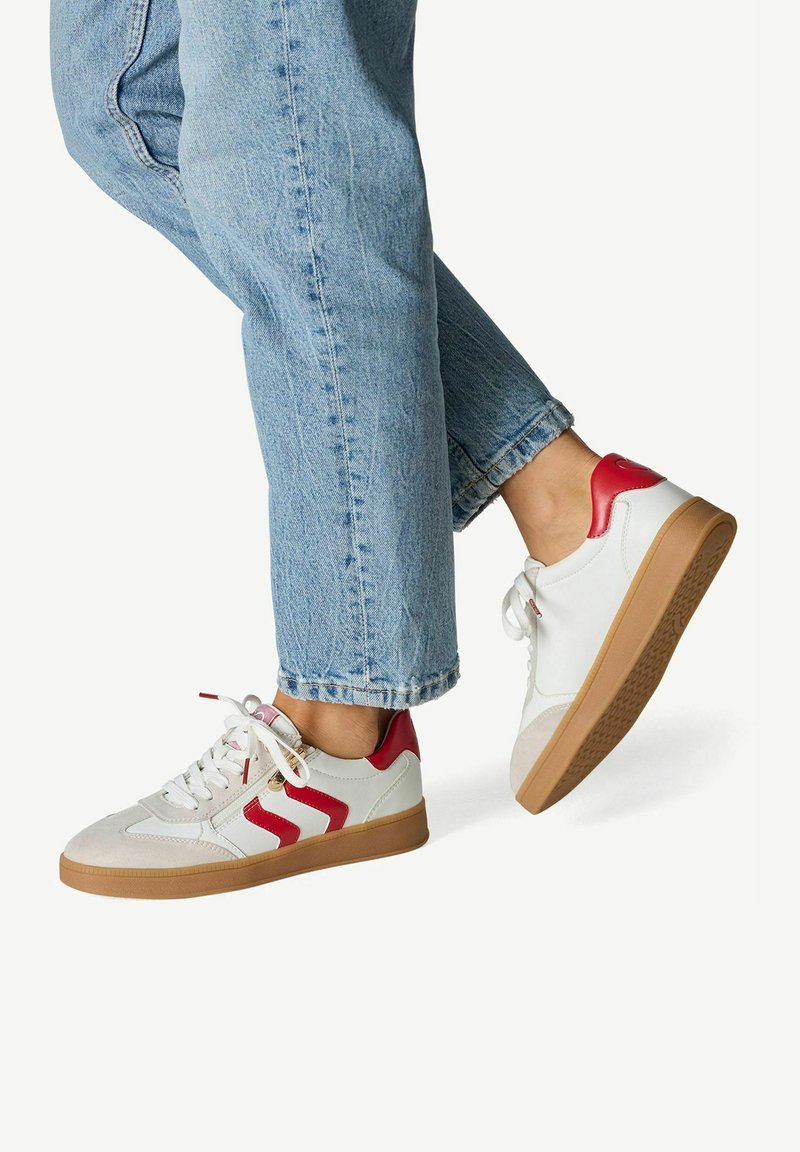 Zapatillas blancas con detalles rojos y suela de goma color caramelo. Presentan una parte superior lisa con un patrón en zigzag y cordones planos. Usadas con vaqueros azules remangados.