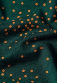 Grön tyg med en mjuk textur, prydd med spridda orangea prickar i olika storlekar, som skapar ett prickigt mönster över ytan.