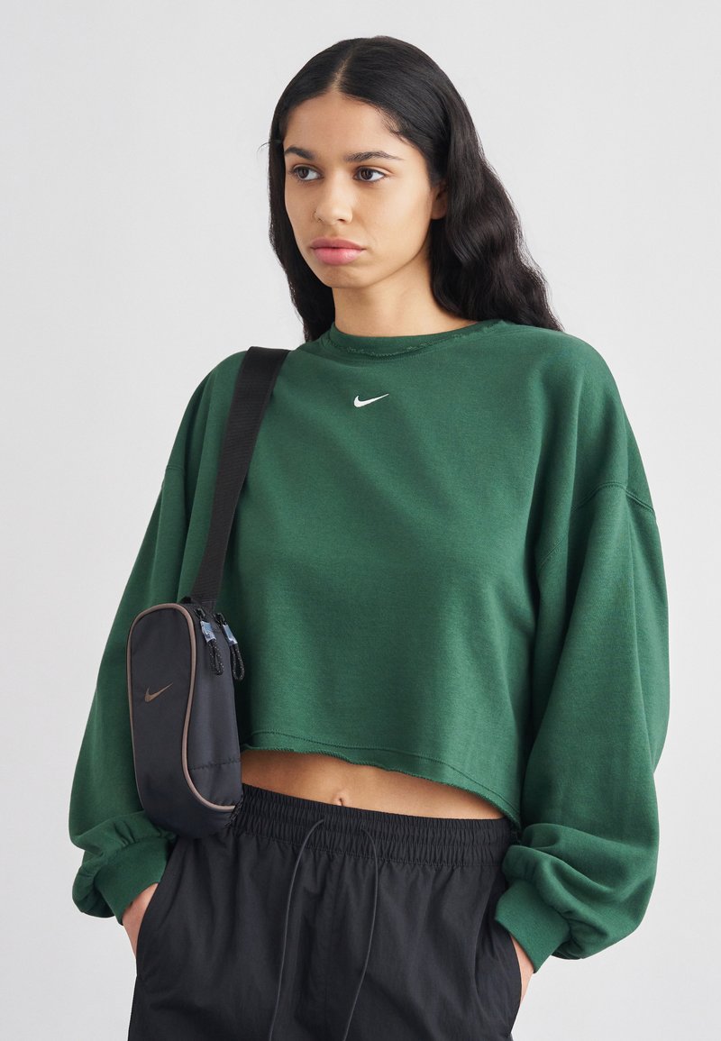 Πράσινο κοντοκομμένο φούτερ με μακριά μανίκια και στρογγυλή λαιμόκοψη, με μικρό λογότυπο Nike. Συνδυάζεται με μαύρα, φαρδιά σορτς και μια μικρή μαύρη τσάντα.