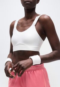 Αθλητικό άτομο που φοράει λευκό αθλητικό σουτιέν, ροζ σορτς Under Armour και λευκά περιβραχιόνια σε απλό ανοιχτό φόντο.