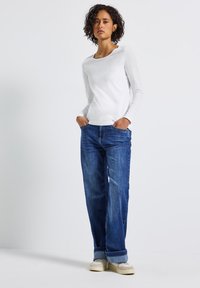 Eine Frau mit kurzen lockigen Haaren, die ein weißes Langarmshirt, blaue hochgekrempelte Jeans und beige Sneakers trägt, steht mit den Händen in den Taschen.