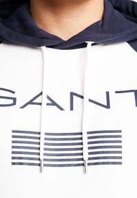 Sudadera blanca con acentos en azul marino, que cuenta con un cordón con extremos de metal y un estampado gráfico de rayas y texto en azul marino en la parte delantera.