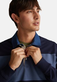 Pánská polo tričko s límcem z modrého denimu, tmavě navy a světle modré barevné bloky a tříknoflíkovou zapínací lištou. Měkký bavlněný materiál.