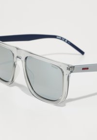 Ochelari de soare din plastic transparent cu lentile gri, accentuați de un brat de culoare albastră închisă și un logo roșu pe braț. Design cu ramă pătrată.