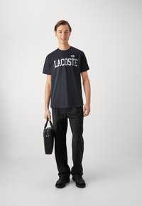 Mornarski kratki rukav majica s natpisom "LACOSTE", uparen s crnim hlačama i cipelama. Drži crnu torbu s dvostrukim ručkama.