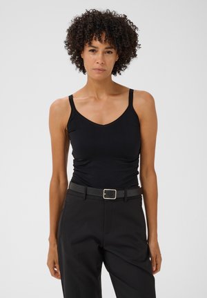 Femme aux cheveux bouclés portant un haut noir sans manches rentré dans un pantalon noir avec une ceinture noire, debout devant un fond uni.