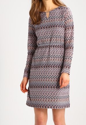 Femme portant une robe longue aux manches longues, longueur genou, avec un motif zigzag multicolore aux tons atténués, debout devant un fond uni.