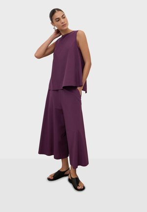 Femme aux cheveux attachés, portant un haut ample sans manches violet et un pantalon large raccourci avec des sandales noires ouvertes, debout sur un fond gris.