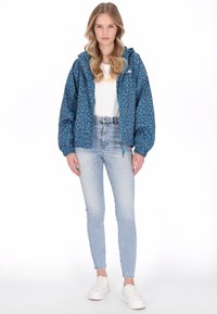 Kék leopárdmintás kapucnis dzseki, rugalmas passzékkal és cipzáras lezárással, fehér póló és világoskék szűk farmer nadrág fölött viselve.