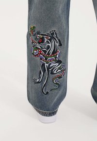 Denim jeans featuring a colorful embroidered design of a panther and a snake on the left leg, with a classic blue wash and cuffed hems.