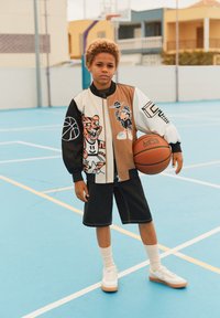 Dreng, der holder en basketball på en blå udendørs bane, iført en flerfarvet jakke med en tegneserie-tiger og -fugl, sorte shorts, hvide sneakers og sokker.