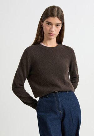 Brown knitted sweater with a round neckline and long sleeves, worn over a light top, paired with high-waisted blue denim trousers.