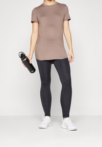 Brun kortärmad sportskjorta, svarta leggings och vita sneakers, hållande en svart vattenflaska. Slät tyg, atletisk passform och enkel design.