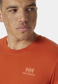 Oranžové bavlněné tričko s kulatým výstřihem. Na levé části hrudi má světlou šedou potisknutou značku "Helly-Hansen". Hladká textura.