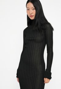 Μαύρο μακρύ μανίκι φόρεμα με σχέδιο καρό, ψηλό γιακά και βολάν στους καρπούς. Φτιαγμένο από διαφανές, υφασμάτιο με υφή. Στενή γραμμή.