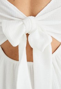 Weißes Kleid aus strukturiertem Stoff, mit einer Schleifen-Detail am Rücken und einem Rückenausschnitt.