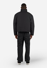Svart polstret jakke med høy krage, vist på en person som har på seg mørke jeans og svarte joggesko, mot en hvit bakgrunn.