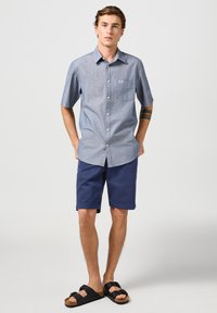 Koszula z krótkim rękawem, zapinana na guziki, w niebiesko-białą kratę, zestawiona z granatowymi szortami. W połączeniu z czarnymi sandałami, charakteryzująca się luźnym krojem i detalem w postaci kieszeni.