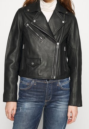 Frau trägt schwarze Leder-Bikerjacke mit Reißverschluss und silbernen Beschlägen über weißem Rollkragenpullover und blauer Jeans, steht vor einfachem Hintergrund.