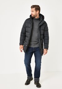 Schwarze Daunenjacke mit Kapuze, mit Reißverschluss vorne, Seitentaschen und gestepptem Muster. Getragen über einem grauen Strickpullover und blauen Jeans.