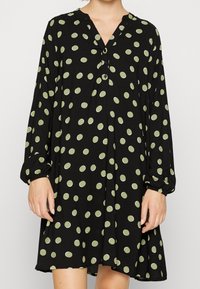 Musta mekko, jossa on pitkät hihat, V-pääntie, vihreitä pallokuvioita ja nappidetaljeja. Kangas näyttää pehmeältä ja kevyeltä, ja mekko on liehuvassa muodossa.