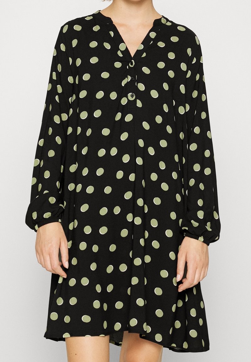 Musta mekko, jossa on pitkät hihat, V-pääntie, vihreitä pallokuvioita ja nappidetaljeja. Kangas näyttää pehmeältä ja kevyeltä, ja mekko on liehuvassa muodossa.