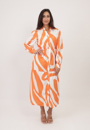Robe à manches longues, boutonnée, aux rayures abstraites orange et blanches, avec une taille cintrée et une longueur midi, fabriquée en tissu lisse. Sandales transparentes.