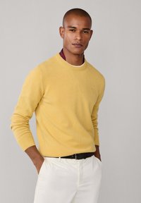 Pull en tricot jaune avec un col rond, des poignets côtelés et un petit logo, associé à un pantalon blanc et une ceinture noire.