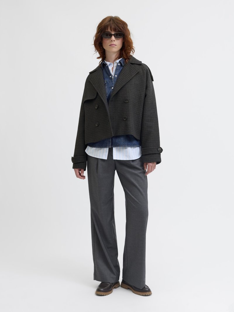Abrigo corto de cuadros negros con botones de doble botonadura, superpuesto sobre una camisa de mezclilla y una camisa blanca rayada, combinado con pantalones anchos grises.