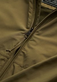 Tissu de veste vert olive avec fermeture éclair noire partiellement ouverte et tirette de fermeture éclair noire avec boucle en cordon.