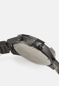 Orologio in metallo nero con cassa rotonda, superficie testurizzata, due pulsanti laterali e un retro minimalista in acciaio inossidabile. Finiture spazzolate e lucidate.
