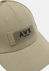 Beżowa czapka baseballowa z teksturowaną, prostokątną naszywką z napisem "AVX" w ciemnoniebieskim kolorze. Materiał to miękka bawełna z widocznymi detalami przeszyć.