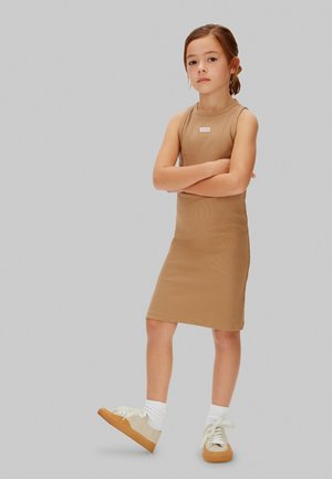 Ung pige med brunt hår i en hestehale iført en ærmeløs beige kjole og hvide sneakers, stående med armene krydset.