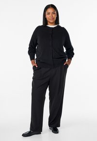 Čierny pletený kardigán so zapínaním na gombíky, v kombinácii s voľnými čiernymi nohavicami. Model stojí s rukami v vreckách, má na sebe čierne topánky.