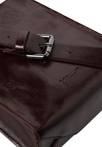 Burgunderfarvet lædertaske med en skinnende sølvspænde, glat overflade og præget brandnavn. Vinklet design med en minimalistisk æstetik.