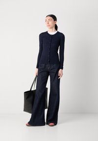 Marinblå ribbad cardigan med knäppning, kombinerad med mörka utsvängda jeans. Modellen håller en svart tote-väska och har på sig rosa och bruna skor.