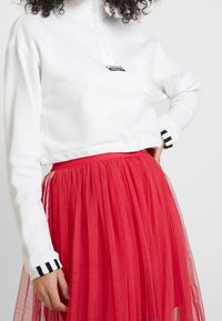 Vit kortärmad sweatshirt med dragkedja och bröstficka, kombinerad med en lager-på-lager röd tyllkjol med svart och vita randiga detaljer.