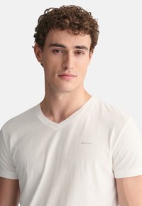 Weißes Baumwoll-T-Shirt mit V-Ausschnitt und kurzen Ärmeln. Dezentes "GANT"-Logo auf der linken Brust. Glatte Textur und figurbetontes Design.