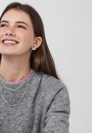 Maglione grigio in lana sovrapposto a un top rosa, con collane d'argento. Orecchino con perla e design floreale all'orecchio sinistro.