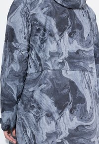 Person som bär en långärmad jacka med ett svart och grått marmorliknande mönster, sedd bakifrån med en del av vänster hand synlig.