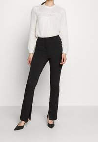 Blusa bianca in tessuto trasparente e texturizzato con dettagli in pizzo sul petto, abbinata a pantaloni neri a vita alta con spacchi laterali.