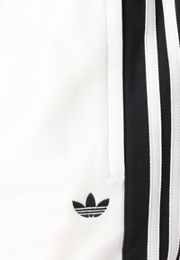 Λευκές αθλητικές βερμούδες με μαύρες λωρίδες πλάγια και κεντητό μαύρο λογότυπο. Φτιαγμένες από υλικό με υφή, αναπνεύσιμο.